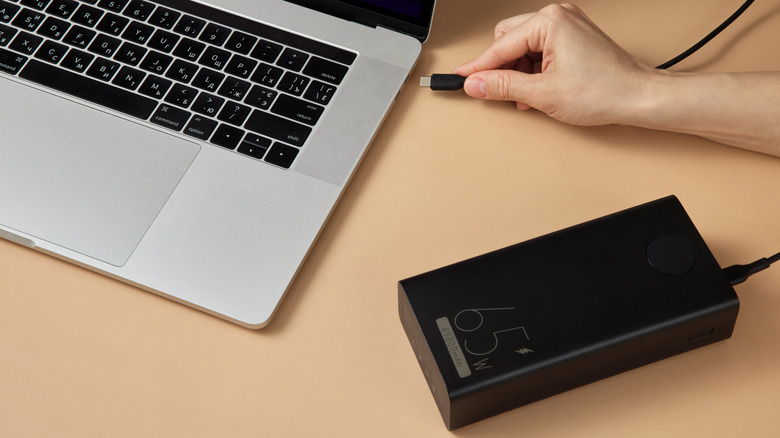 Hand plugging a 65W power bank into a laptop charging port.
