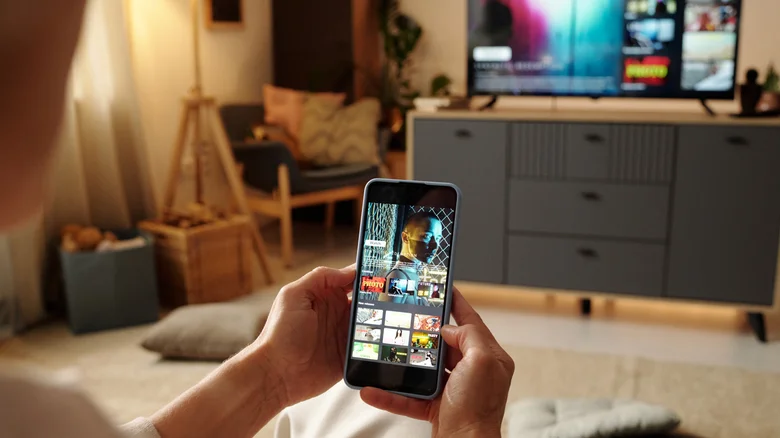 Hands holding a cell phone showing a movie with the same image on the TV.