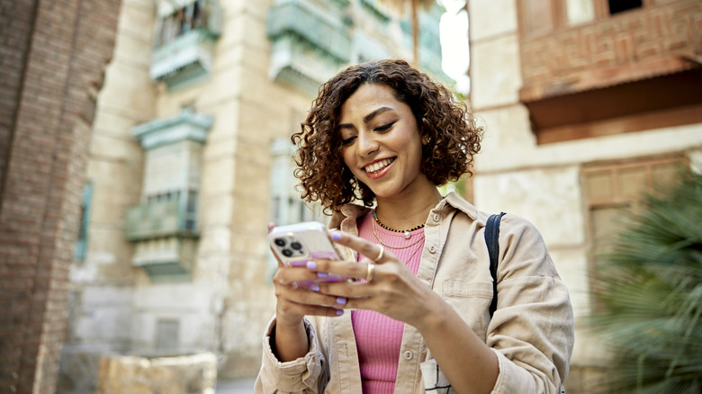 A woman in a pink shirt and tan jacket uses her phone as she walks away from a brick apartment building