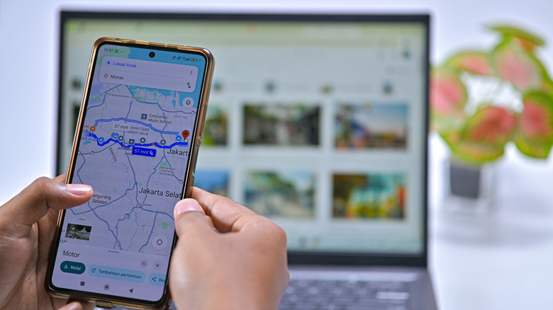 Hands holding a smartphone showing Google Maps navigation, with a blurred laptop in the background.