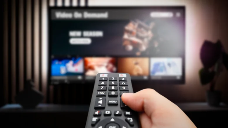 A hand holding a remote in the foreground with a TV blurred out in the background.