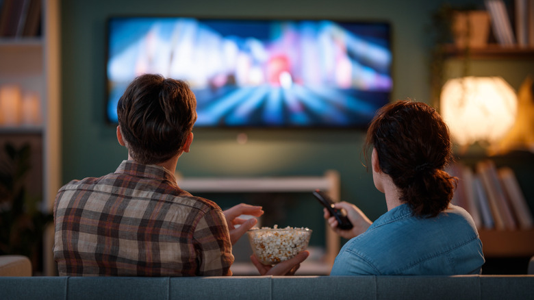 The back of a couple watching TV, the woman holding the remote and the man a bowl of popcorn.