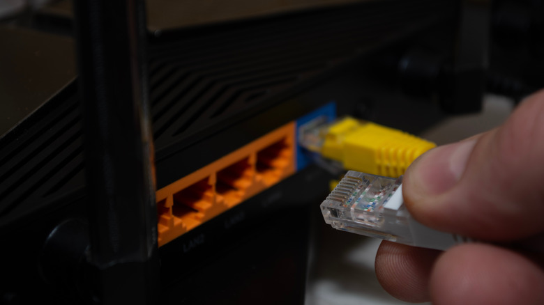 A hand holding a clear Ethernet connector beside a black router with orange LAN ports and a yellow cable connected to a blue port.