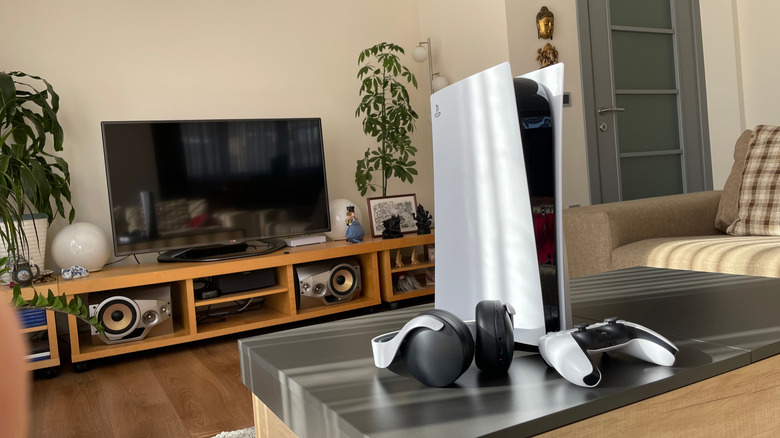 A PS5, next to a set of headphones and a controller, with a TV on an entertainment center in the background.