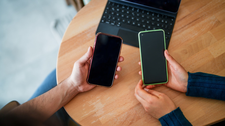 Two people holding phones with black screens at a wooden table.