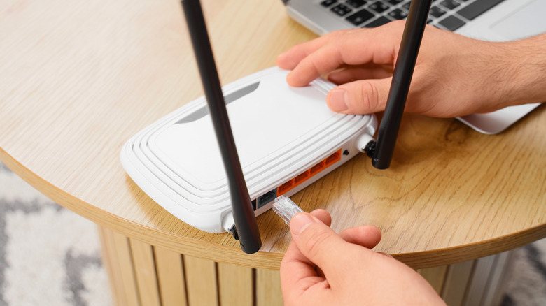 Person plugging an Ethernet cable into a wireless router, near a laptop on a table.
