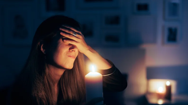 A woman slaps her forehead while holding a candle in a lightless room.