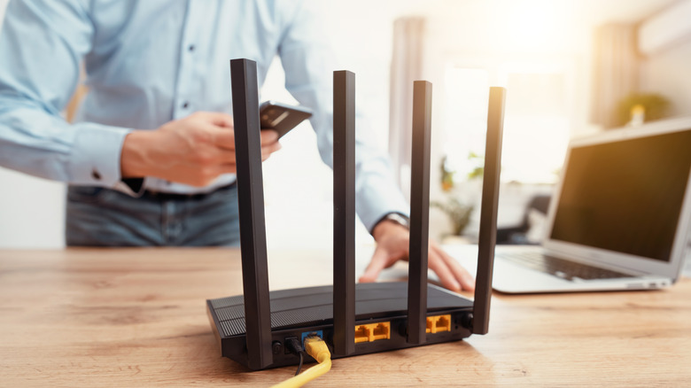 The rear view of a Wi-Fi router, showing antennas at the back, with a man holding a phone close to a laptop in the rear.