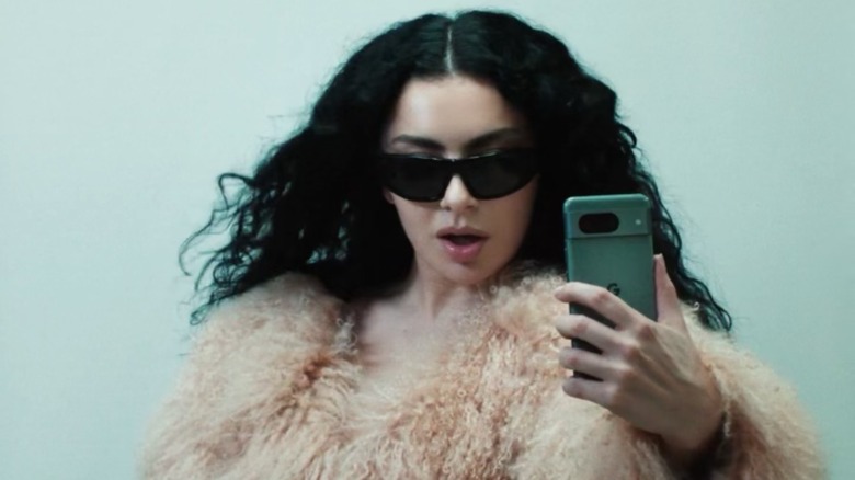 Pop star Charli XCX, in a puffy jacket and shades, poses as she takes a photo with a green Android phone.