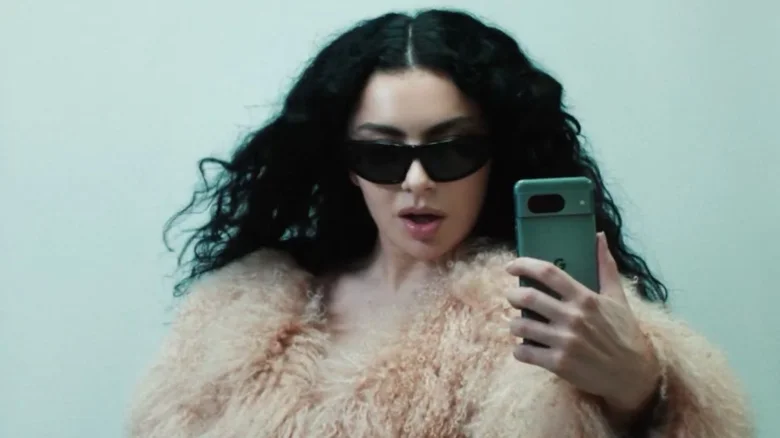 Pop star Charli XCX, in a puffy jacket and shades, poses as she takes a photo with a green Android phone.