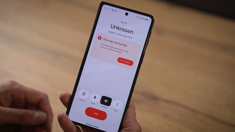 A man holding a phone during a call from an unknown number receives a red scam notification stating "This may not be Dan," with the words :someone may be pretending to call from your contact's number" written beneath it.