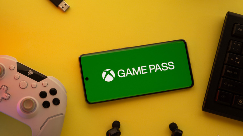A top view of a phone screen with the Xbox Game Pass logo, a controller, earbuds and a keyboard on a yellow surface.
