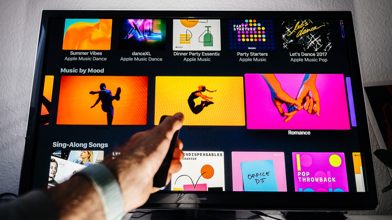 A hand holding a remote in front of a TV showing music album covers.