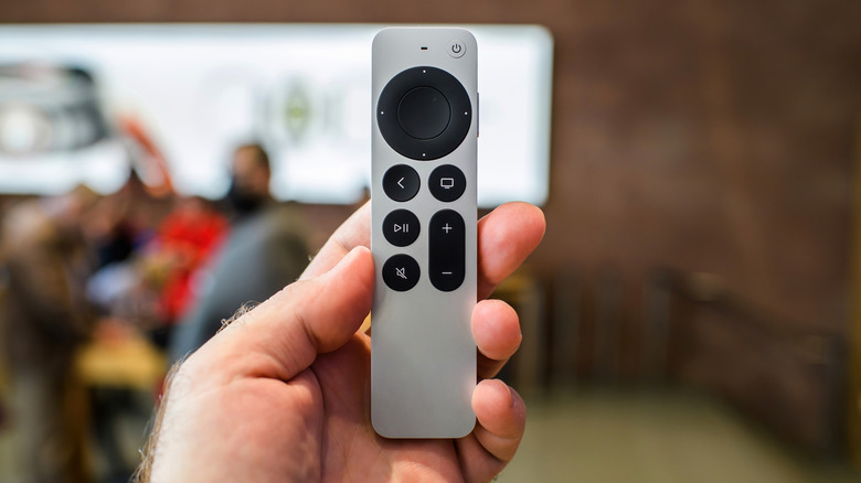 A hand holding up a new Apple TV Siri remote with faded out people in the background.