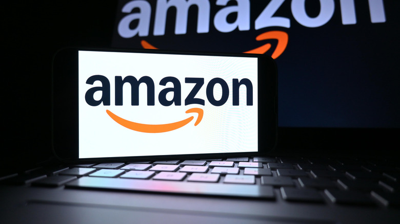 Phone screen displaying the Amazon logo with an open laptop displaying the same in the background.