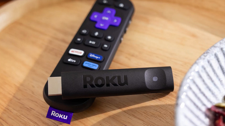 A Roku streaming stick sits across Roku's iconic remote atop a wooden table.