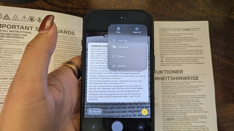 A hand holding an iPhone 17 Pro up to an instruction manual showing the various options for Live Text.