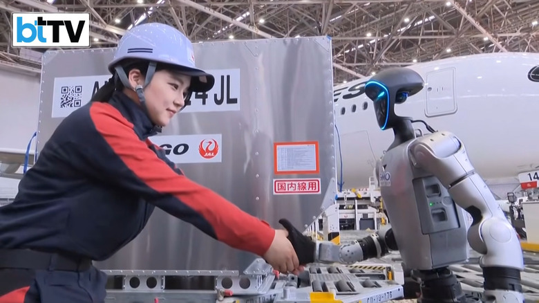 A worker with a hardhat shaking hands with a humanoid robot. Planes in background.