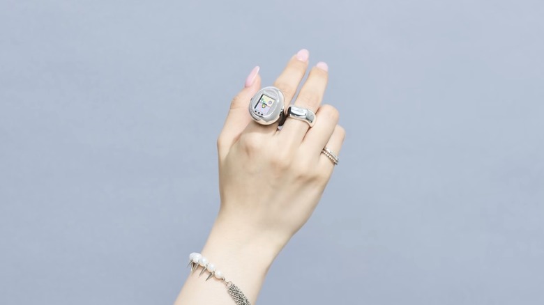 A hand sporting a silver and black Tamagotchi ring on the index finger, alongside normal silver rings on the middle and ring fingers, is held up against a light blue-gray backdrop.