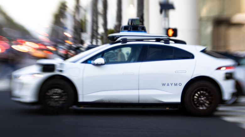 White Waymo self-driving vehicle moving through a city intersection with its roof-mounted autonomous driving sensors visible.