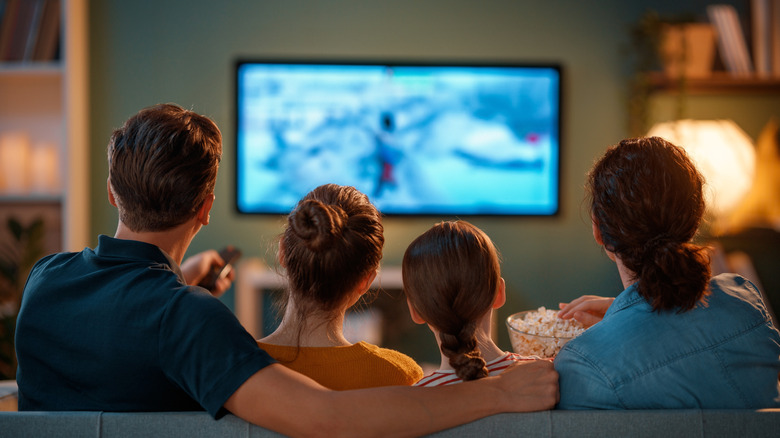 A family seated on a couch watching TV and eating popcorn.