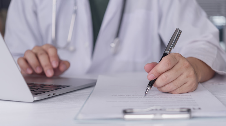 A doctor using a pen to write on a clipboard while using a laptop.