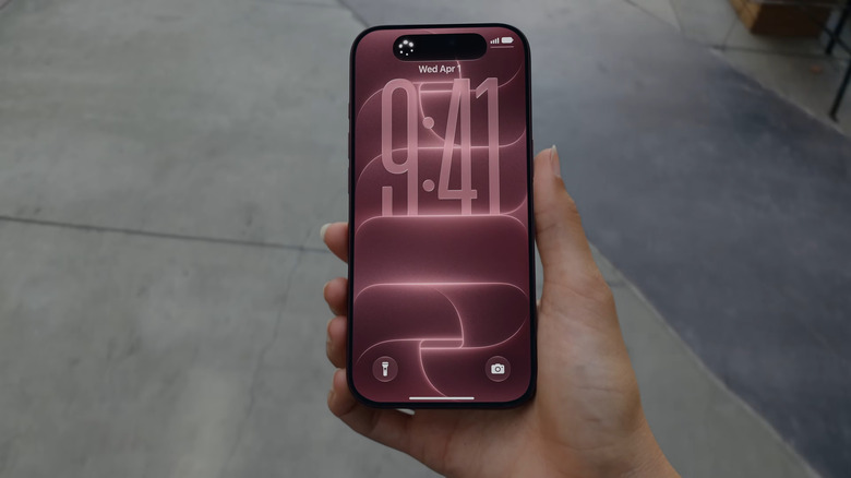 A hand holding the iPhone 18 Pro, with its (burgundy wallpaper) lock screen showing.
