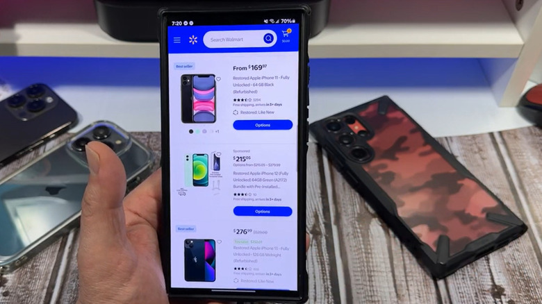 A man holds a phone up, showing the Walmart Marketplace app and several used iPhones available for purchase.