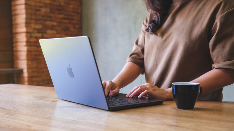 A woman standing up working on a MacBook with a coffee mug beside her.