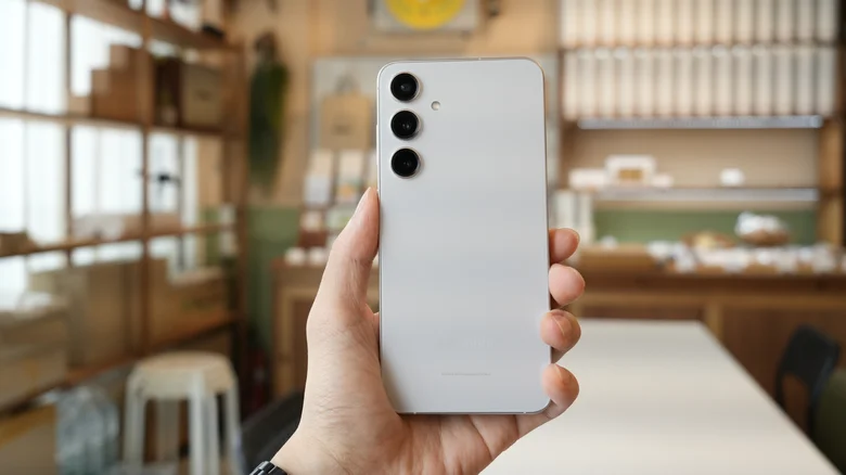 A person holding a silver Samsung Galaxy phone with three rear cameras, in a softly blurred indoor setting.