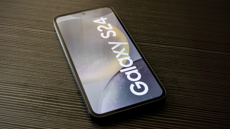 Samsung Galaxy S24 smartphone lying face-up on a dark textured surface, with the Galaxy S24 name displayed on the screen.