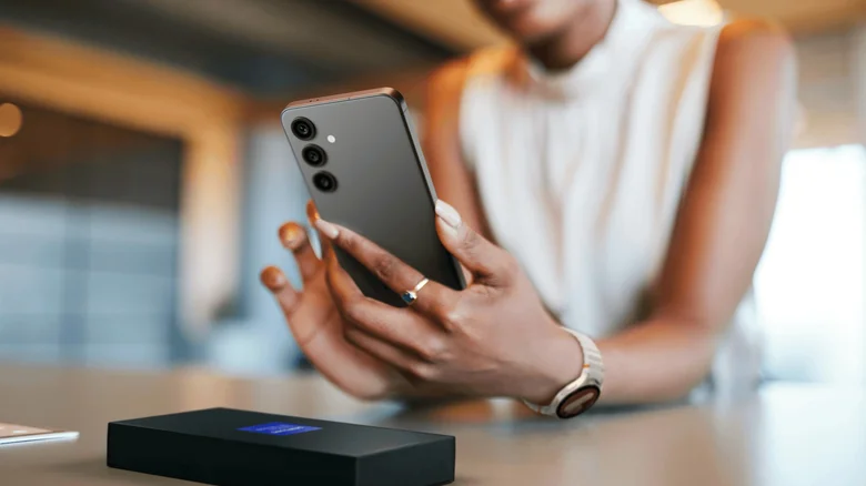 A person holding and using a dark Samsung Galaxy phone above a table, with the phone's rear cameras visible.