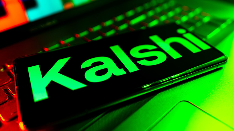 Photo of a smartphone displaying the Kalshi logo. It sits on a laptop keyboard and is illuminated by a green light.