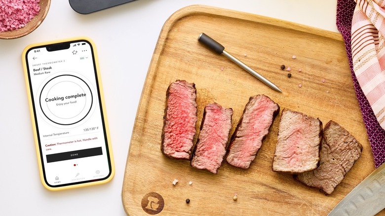 Top-down photo of a KitchenAid Smart Thermometer next to slices of cooked meats at different donenesses on a wooden cutting board. A smartphone to the left reads 