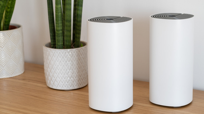 Two white mesh Wi-Fi nodes stand beside green plants in white pots atop a brown table.