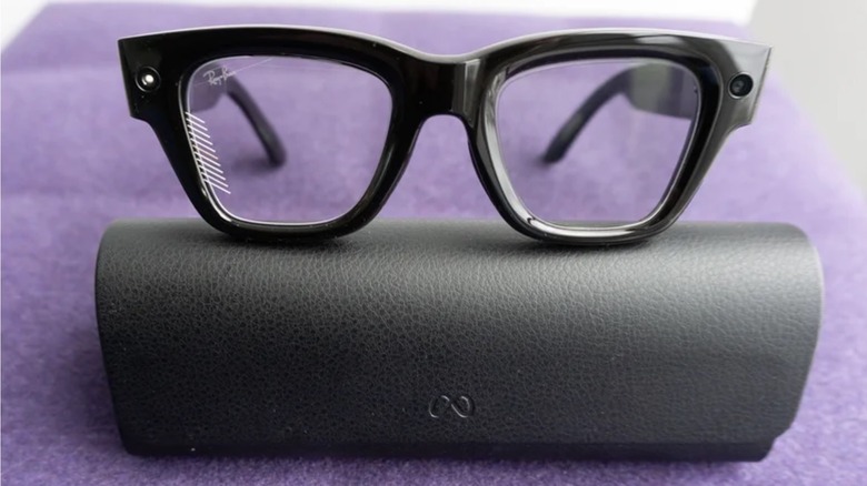 Photo of the Meta Ray-Ban Display smart glasses. Heavy black-framed smart glasses and the accompanying black case arranged on a purple surface.