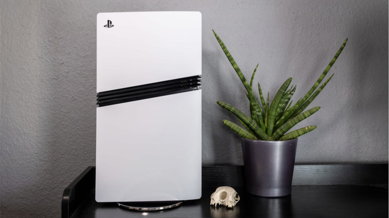 Photo of the PlayStation 5. A white gaming console stood vertically next to a green plant in a silver pot.