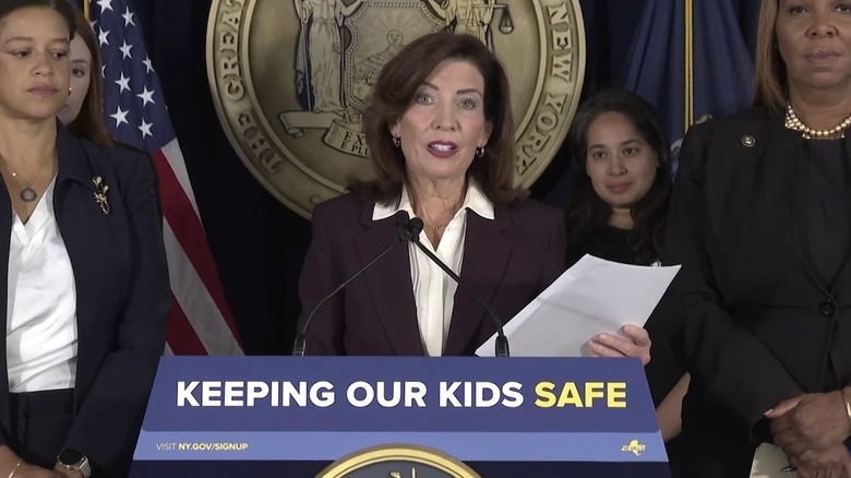 NY Governor Kathy Hochul surrounded by others as she stands behind a lectern with a sign that reads 