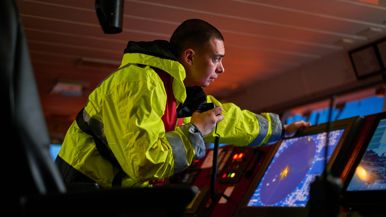 A sea captain in a bright yellow jacket leans over his communication and radar systems, holding a radio microphone.