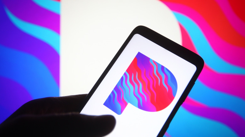 A hand holds up a phone with the Pandora Music logo on its screen, seemingly having ripped it from the Pandora logo in the background.