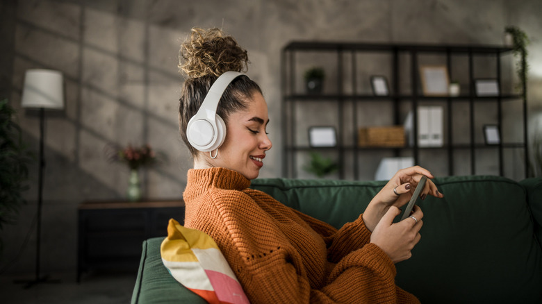 A woman wearing white headphones and an orange sweater smiles as she chooses music on her phone.