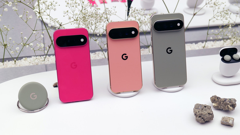 The Google Pixel 10, 10 Pro, and 10 Pro XL standing upright on a white table among a backdrop of white flowers.