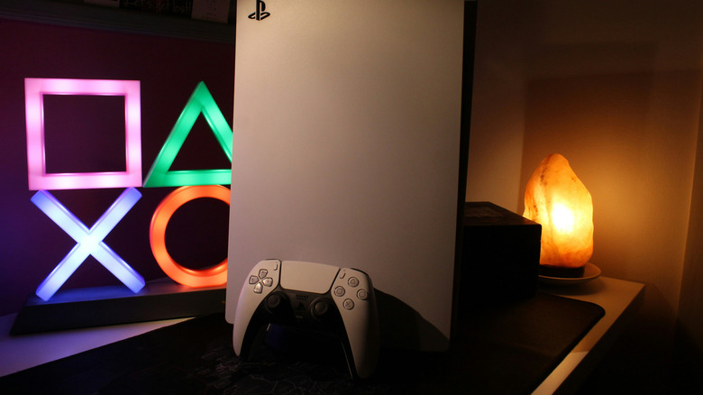PlayStation 5 and controller next to a neon sign of the brand's cross, circle, triangle and square icons.