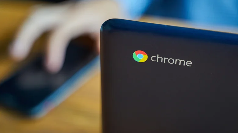 A Chromebook with the logo facing the camera as someone uses a phone on a table in the background.