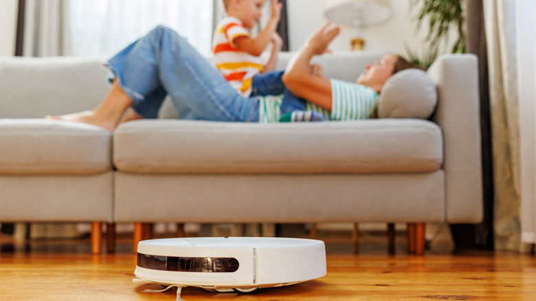 Robot vacuum in the foreground; a mother playing with her child on the couch in the background.
