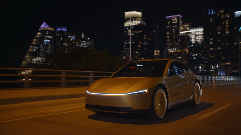 A Tesla Cybercab drives along a bridge at night, a bright cityscape in the background.