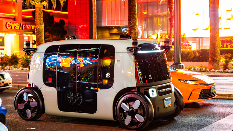 A boxy white Zoox robotaxi oulls up to a stoplight at night, surrounded by other vehicles.