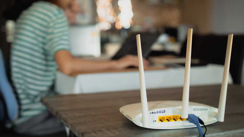 A Wi-Fi router with an Ethernet cable plugged in, with a person working on a laptop blurred in the background.
