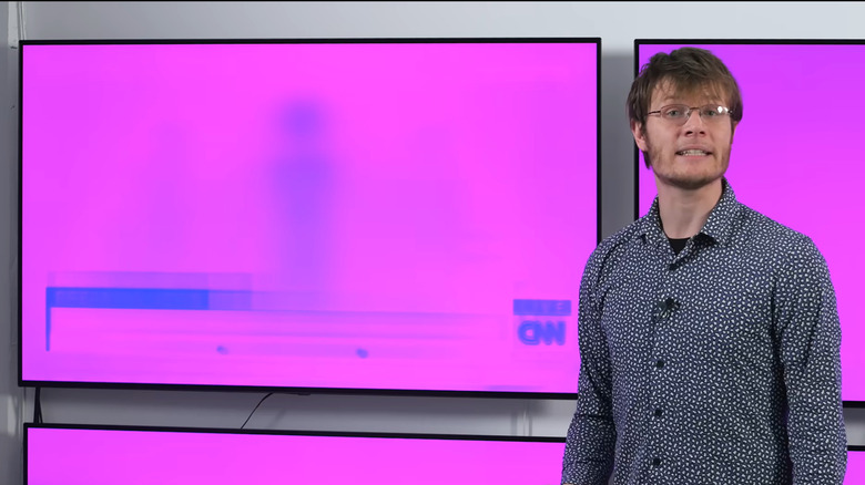 A man in a dark shirt and glasses stands besides a magenta television with blue burn-in spots showing a CNN logo and news ticker.