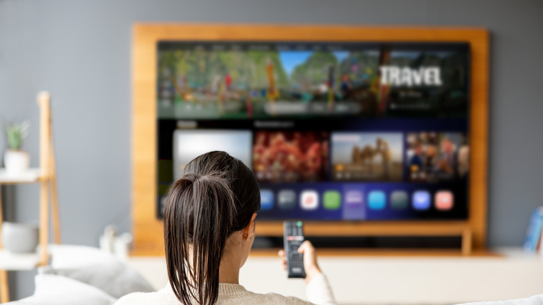 Woman sitting at home, point a remote control at the TV while watching it.